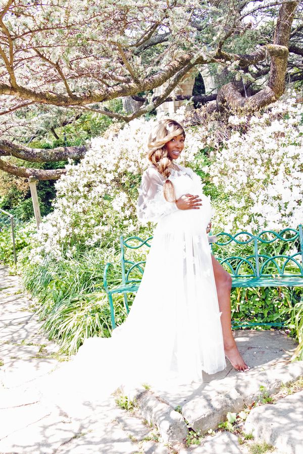 Woman in a white dress standing in a garden with blooming trees. Vote Ruby Lynn – Baby of the Year 2025 campaign