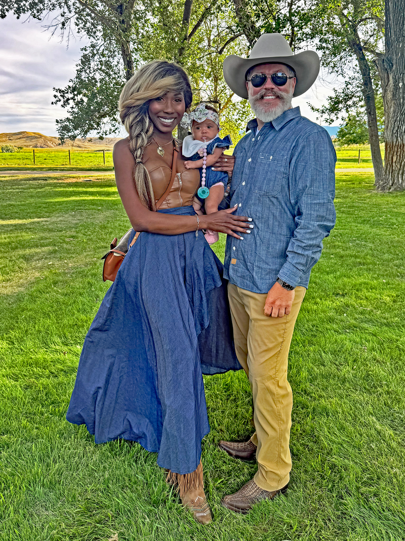 Family of three outdoors in a grassy area with trees and a fence in the background. Vote Ruby Lynn – Baby of the Year 2025 campaign